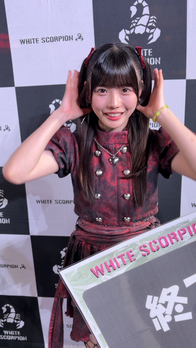 A young woman with dark hair in pigtails and a red and black outfit poses with her hands near her head in front of a 'WHITE SCORPION' checkered backdrop, holding a sign reading 'WHITE SCORPION'.