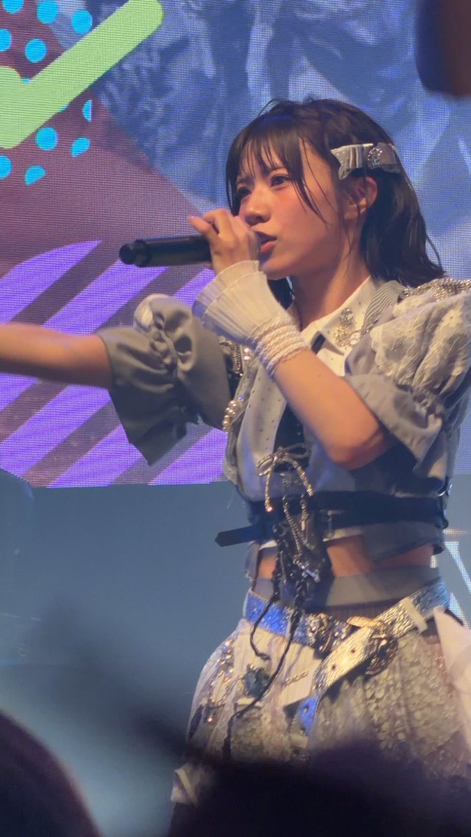 A young female performer with dark hair sings into a microphone while wearing an ornate silver and blue costume with layered sleeves and beaded arm accessories, performing against a colorful LED screen backdrop.