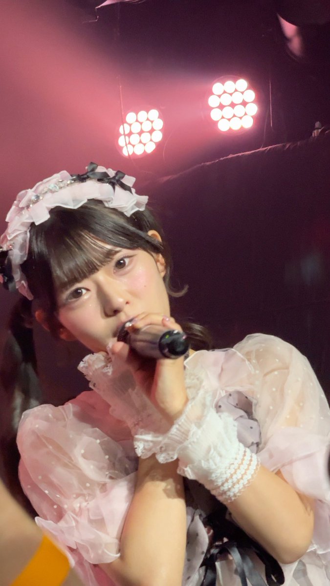 A young woman performing on stage with a microphone, wearing a frilly pink and white outfit with a decorative headpiece, illuminated by stage lights against a pink-hued background.