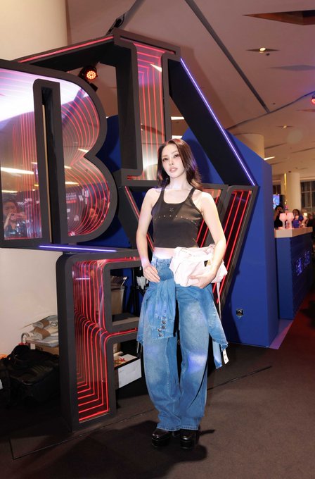 A stylish attendee poses confidently in a black crop top and oversized jeans with a second denim piece tied at her waist, standing in front of the illuminated "BIFW" (Bangkok International Fashion Week) signage. The image captures a moment from the MERGE brand's showcase at #BIFW2026, featuring lookkaew kamollak alongside the event's signature neon backdrop.