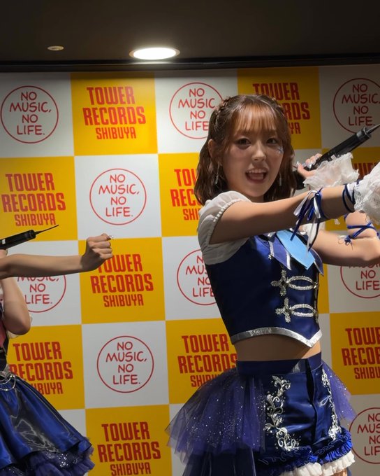 A young woman in a blue and white outfit performs on stage in front of a backdrop displaying 'TOWER RECORDS SHIBUYA' and 'NO MUSIC, NO LIFE' logos.