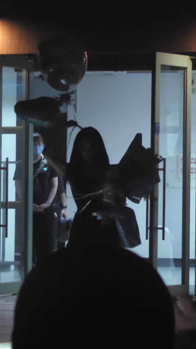 A dark, shadowy image showing silhouetted balloons and a person in dark clothing entering through glass doors, with security staff visible in the background.