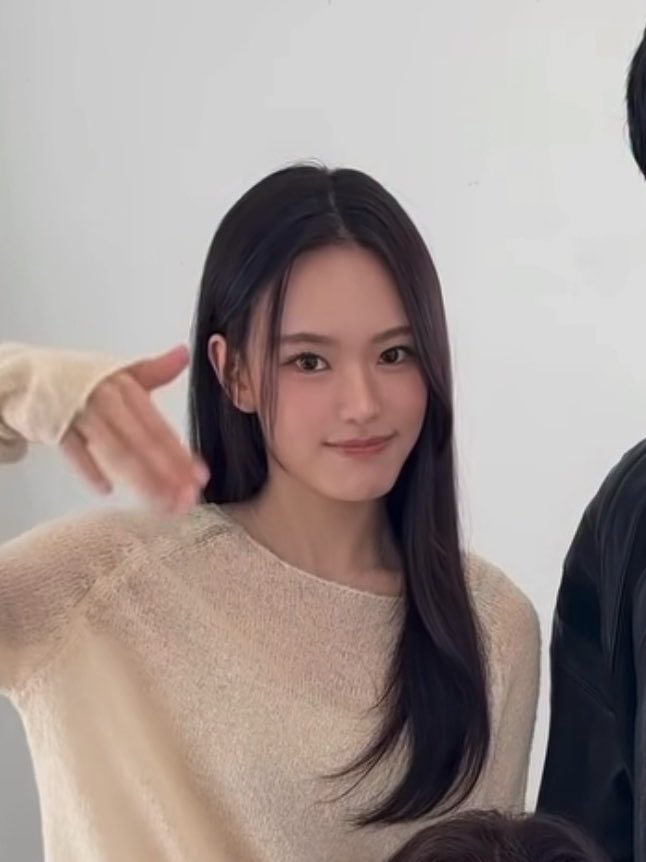 A young woman with long, dark hair wearing a fuzzy beige sweater looks softly at the camera, with another person's hand and shoulder partially visible nearby.