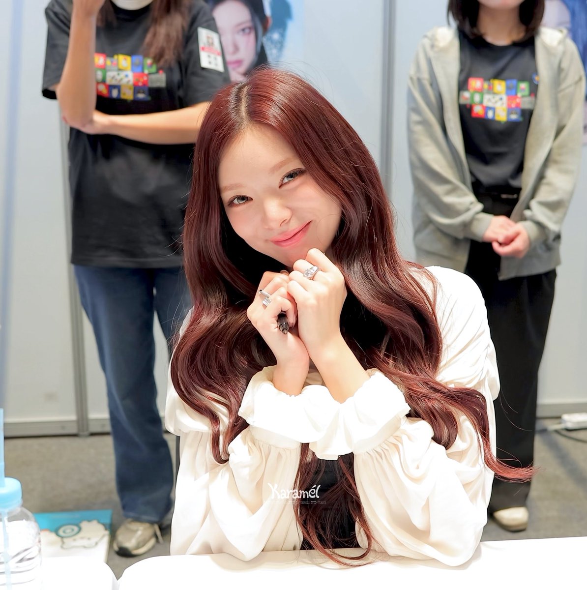A young woman with long wavy auburn hair smiles softly at the camera with her hands clasped near her chin, wearing a white blouse in what appears to be a fan event or signing with staff in the background.