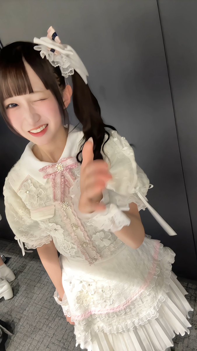 A young woman in an elaborate white and pink frilly Lolita dress winks playfully at the camera with a bright smile. The image captures a candid, joyful moment—likely a fan hoping their favorite performer would wish them happiness on their birthday.