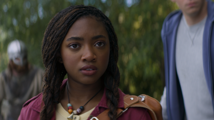 A close-up of a young Black woman with braided hair wearing a beaded necklace, looking concerned, with a masked figure visible in the background.