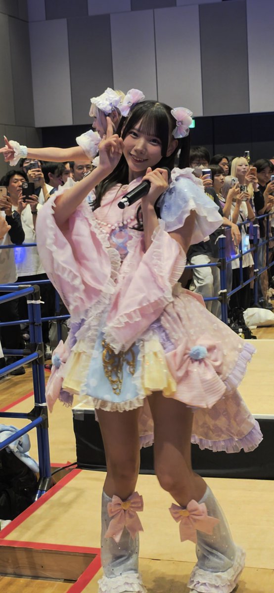 A young woman in a colorful pastel frilly idol outfit with a pearl necklace holds a microphone and smiles while performing on stage. Behind her, fans lean over a blue railing taking photos with their phones.