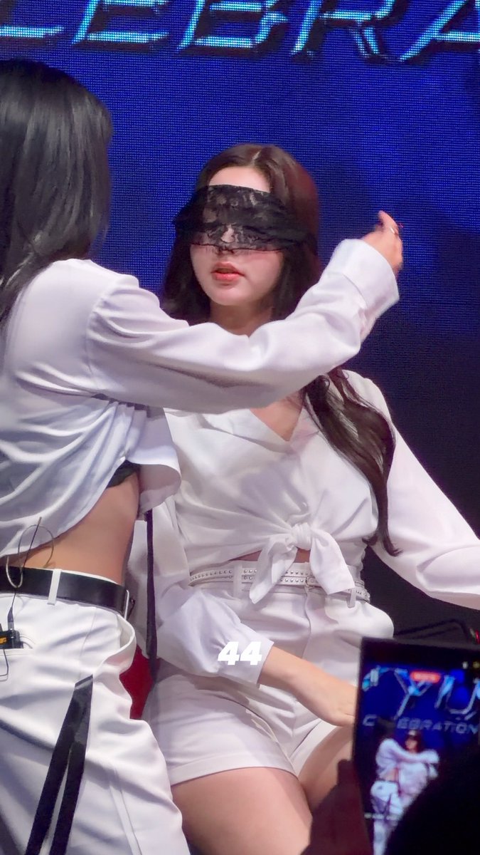 Two performers in white outfits appear on stage; one is blindfolded with lace while the other reaches toward her, with the number "44" visible on screen.