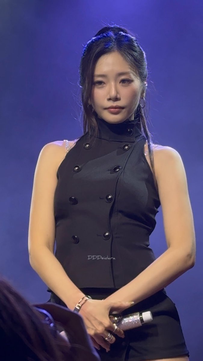 A young woman with long dark hair wearing a black sleeveless double-breasted top stands on a stage holding a microphone, with a watermark reading "DDDcidora".