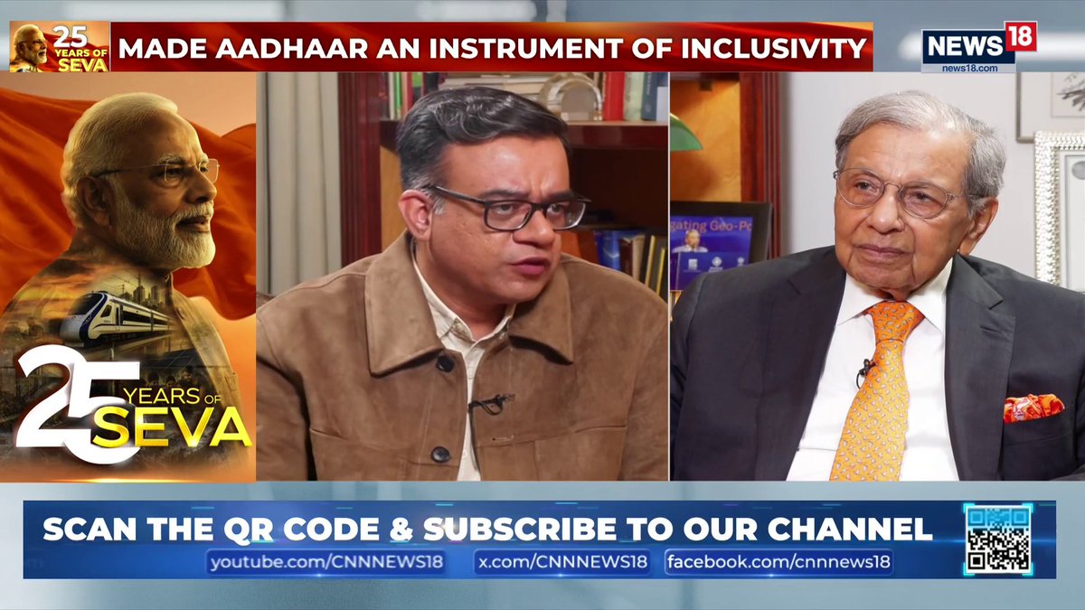 A news broadcast from News18 displays the headline "MADE AADHAAR AN INSTRUMENT OF INCLUSIVITY" alongside a "25 YEARS OF SEVA" graphic featuring PM Modi, while two men in an interview setting—presumably economist N.K. Singh and host Aman Sharma—discuss India's economy and the Viksit Bharat Campaign. A banner at the bottom prompts viewers to scan a QR code and subscribe via YouTube, X, and Facebook for CNN News18.