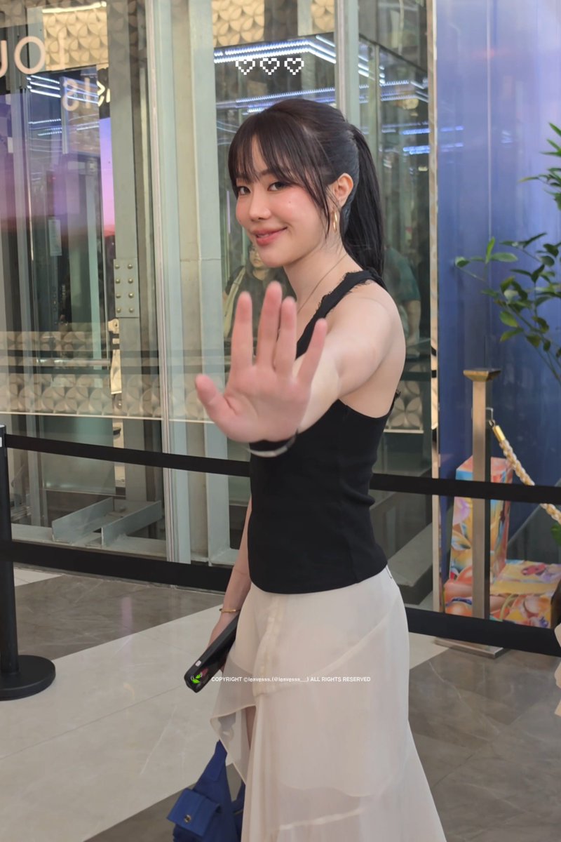 A young woman with a ponytail wearing a black top and white skirt waves at the camera in what appears to be a shopping mall, with a watermark reading 'COPYRIGHT ©lovepsss_(o)lovepsss__ALL RIGHTS RESERVED'.