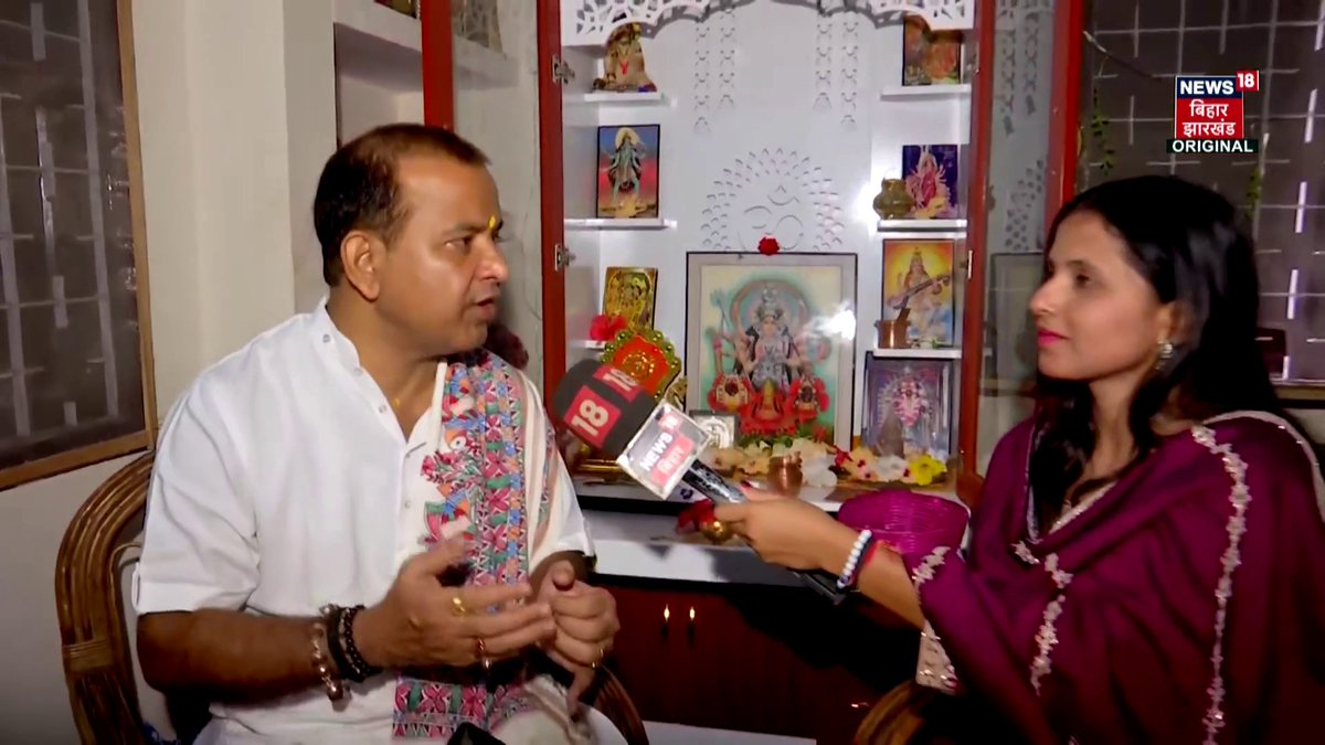 A News18 Bihar Jharkhand interview where a female reporter in a magenta outfit holds a microphone toward a man in a white embroidered vest sitting in front of a Hindu prayer shrine with multiple deities.