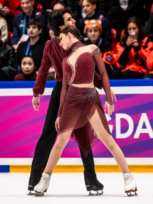 Two figure skaters in coordinated maroon outfits perform a dance routine on the ice. They are surrounded by an audience, with some people dressed in orange jackets. The arena has a large sign with "Gdańsk" visible in the background.