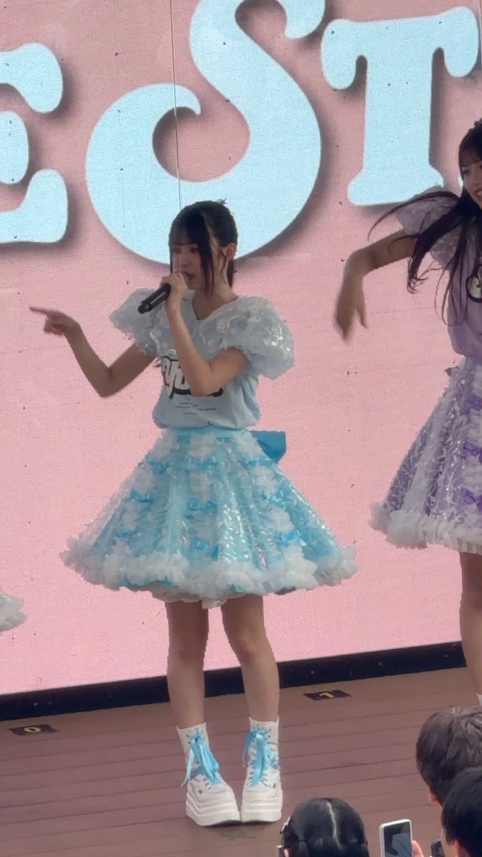 A young female performer in a frilly blue and white idol dress sings into a microphone on stage with a pink backdrop.