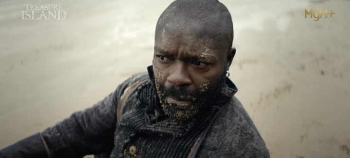 Cinematic still from the MGM+ show 'Treasure Island' showing a close-up of a bearded man with dirt on his face wearing a dark coat, looking intently toward the camera.