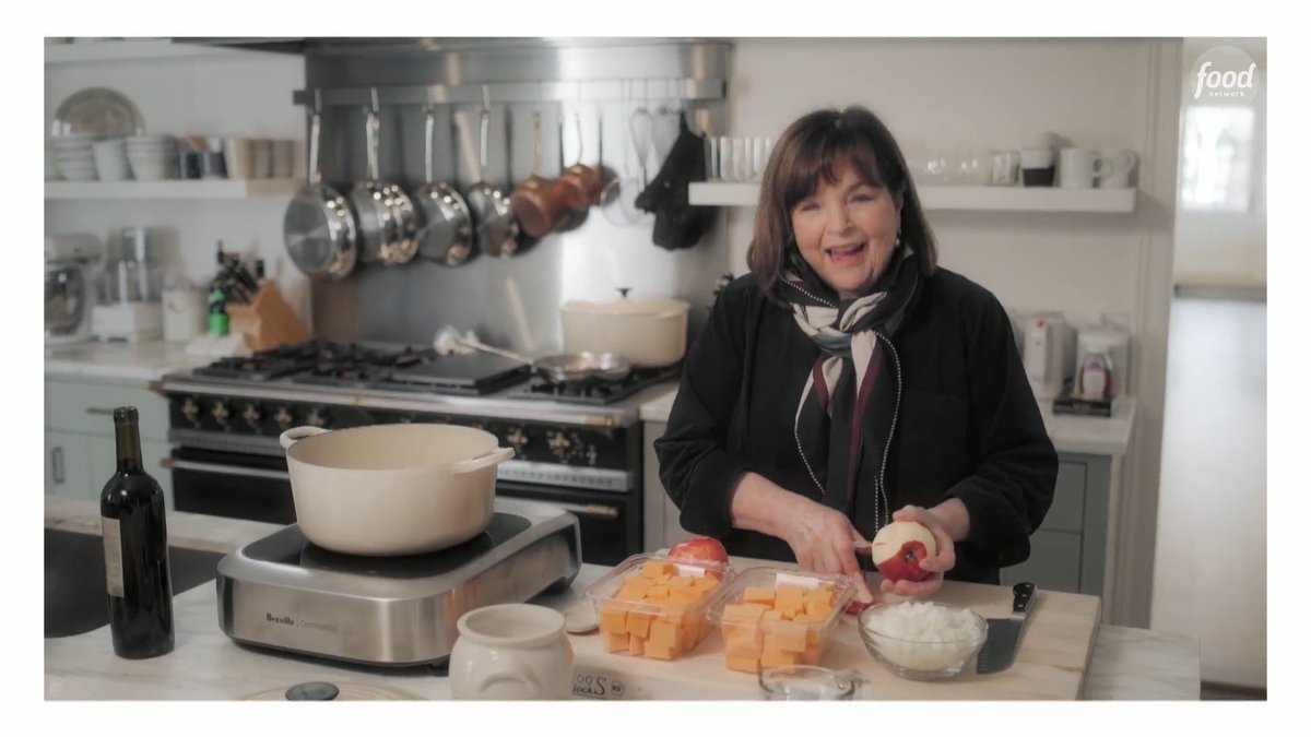 A woman with dark hair and a scarf stands in a kitchen setting preparing food, with a large pot on a stove, chopped butternut squash, onions, and an apple on the counter, with a Food Network logo visible.