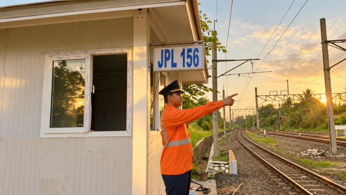 A railway worker in an orange uniform and cap stands beside a small white building labeled 'JPL 156', pointing down a track during sunset.