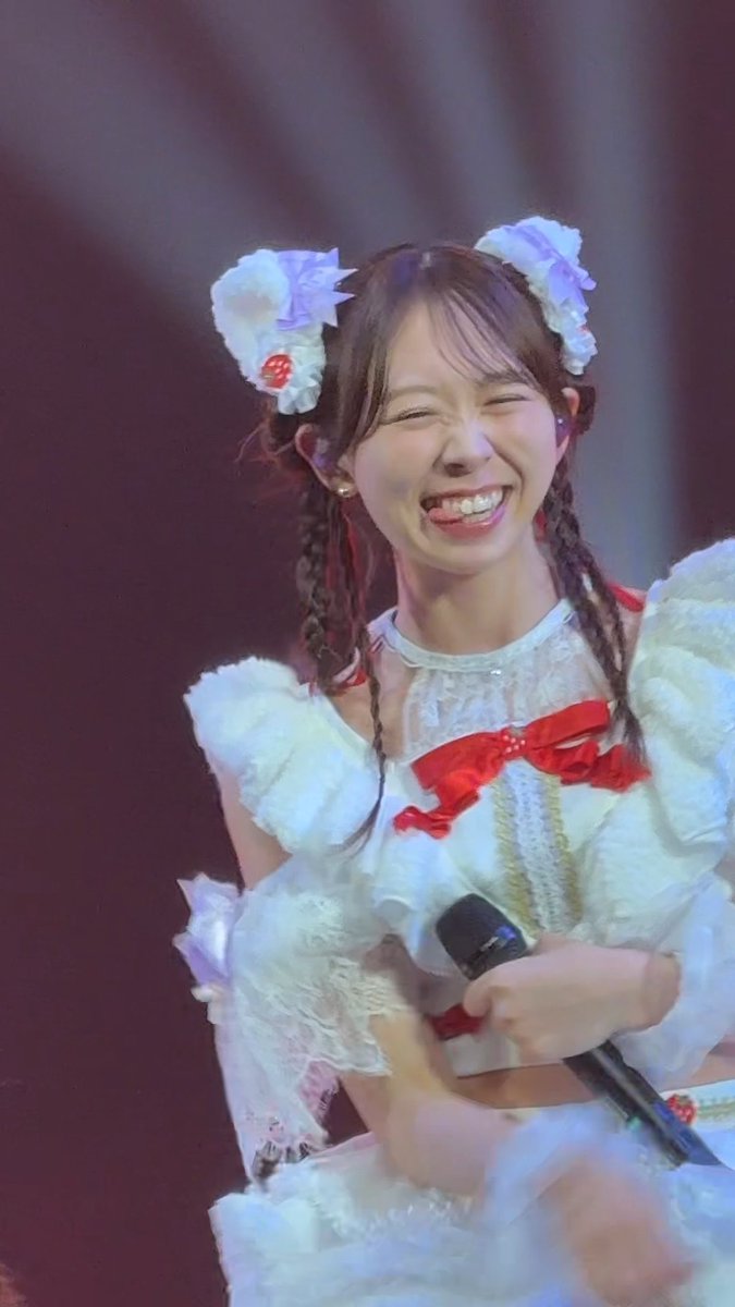A young woman on stage wearing a white frilly idol costume with red bows and matching hair accessories, smiling brightly with her eyes closed while holding a microphone.