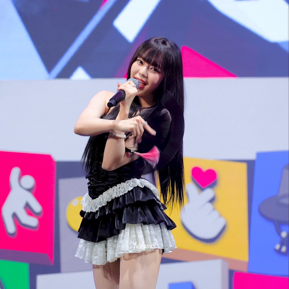 A performer with long black hair and bangs is shown on stage holding a microphone, wearing a black sleeveless top and a tiered black and white skirt.