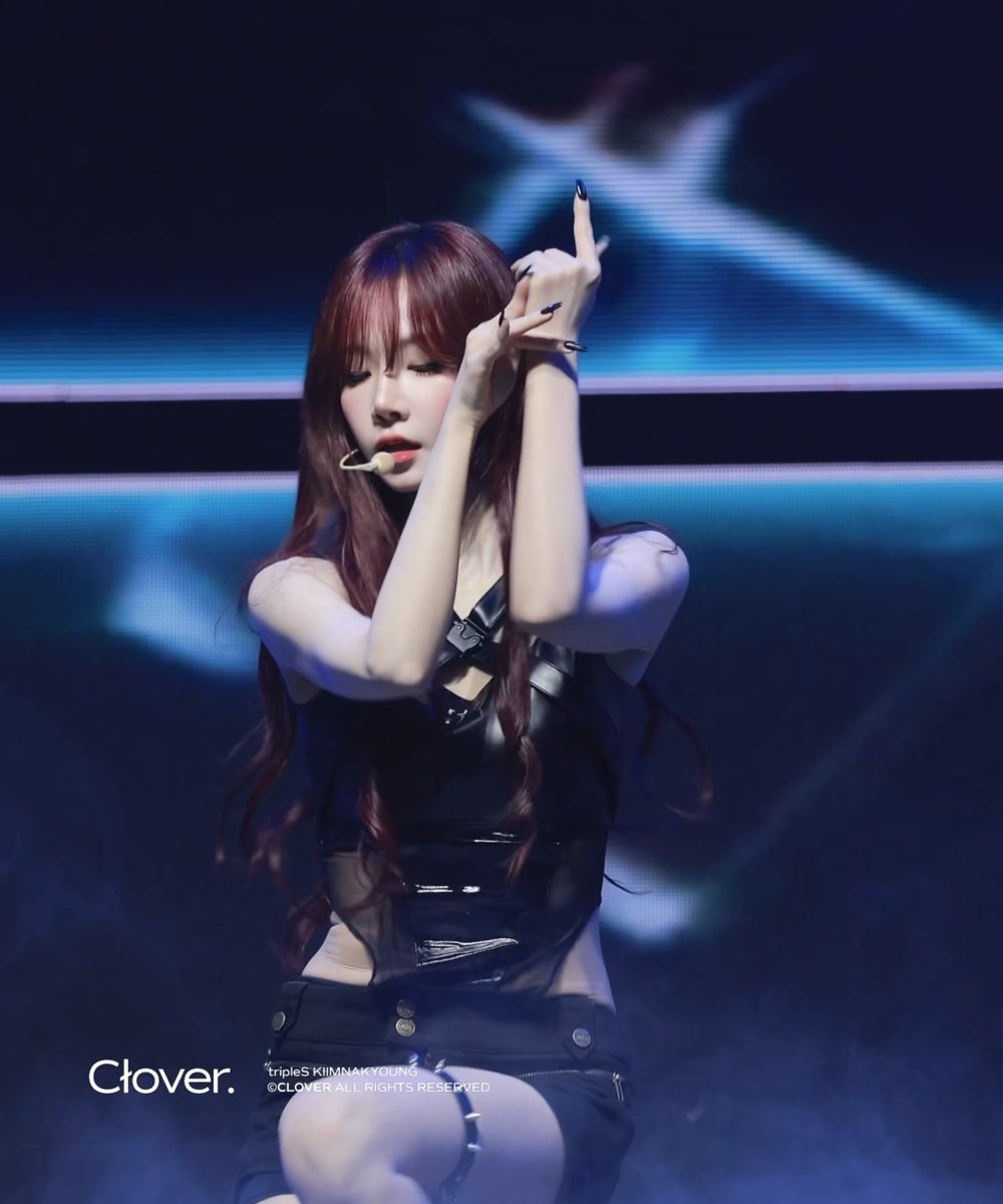 A performer with long reddish-brown hair poses on stage with arms crossed near her face, wearing a black vinyl-style top with buckles and harness details, with the watermark "Clover." and "tripleS KIMNAKYOUNG ©CLOVER ALL RIGHTS RESERVED" visible.