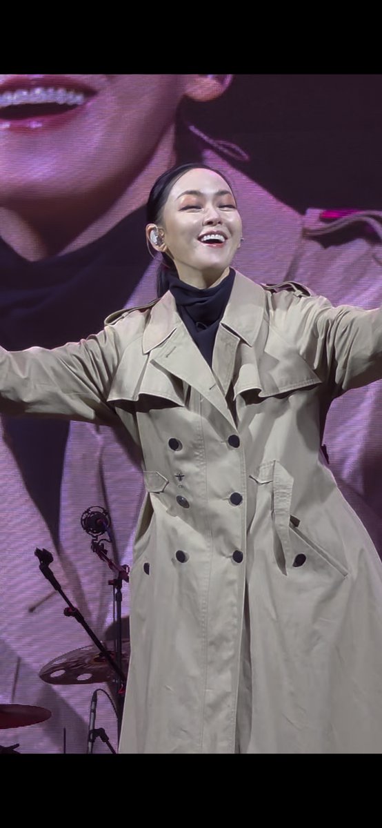 A woman with dark hair pulled back performs on stage, wearing a beige trench coat and smiling broadly with her eyes closed, with drums visible in the background.