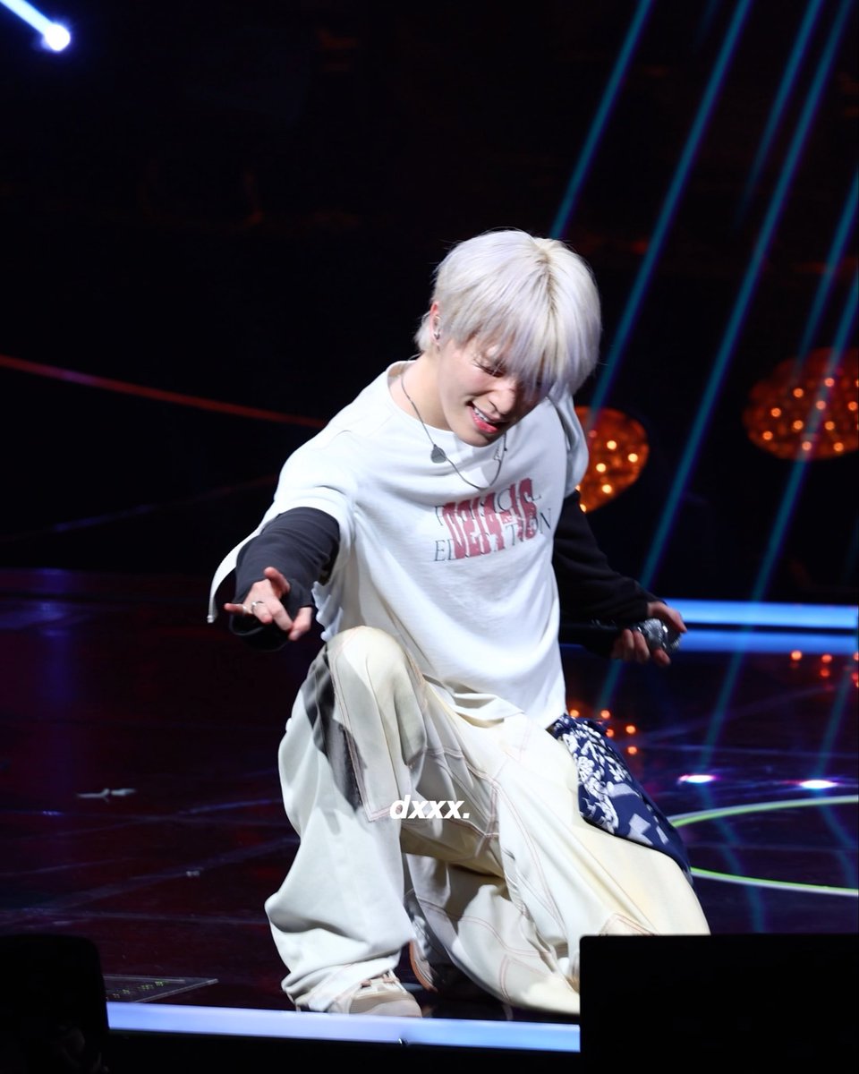 A performer with platinum blonde hair, wearing a white graphic tee and cream-colored pants, crouches on stage while smiling and reaching toward the audience, with a microphone in hand. The watermark "dxxx." appears on the image, and the post identifies the performer as DINO (디노), with the number "261010" likely referencing a date or event code.