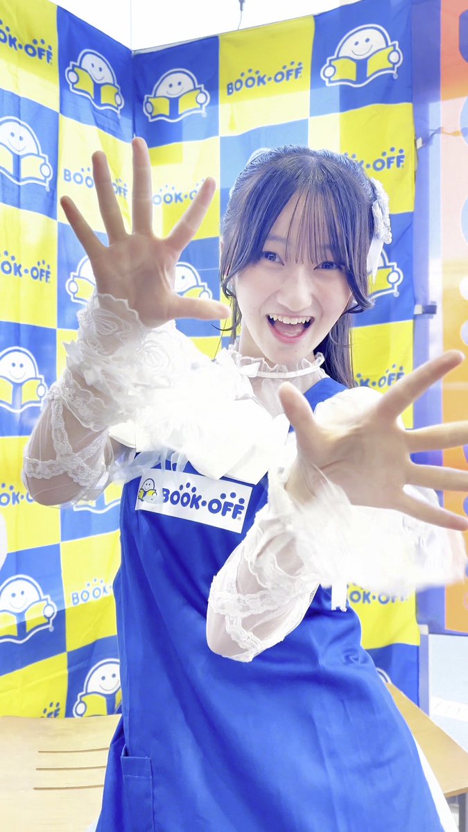 A cheerful young woman wearing a 'BOOK-OFF' branded blue apron over a frilly white top, posing with her hands outstretched in front of a yellow and blue checkered 'BOOK-OFF' backdrop.