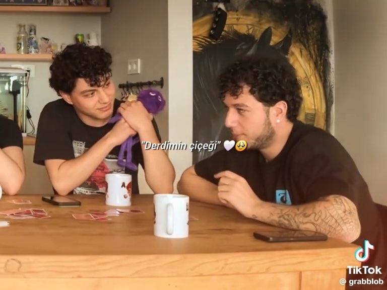 Two curly-haired young men sit at a wooden table with mugs; one holds a purple plush toy and stares at the other, captioned 'Derdimin çiçeği 🤍🥲' in a TikTok video.