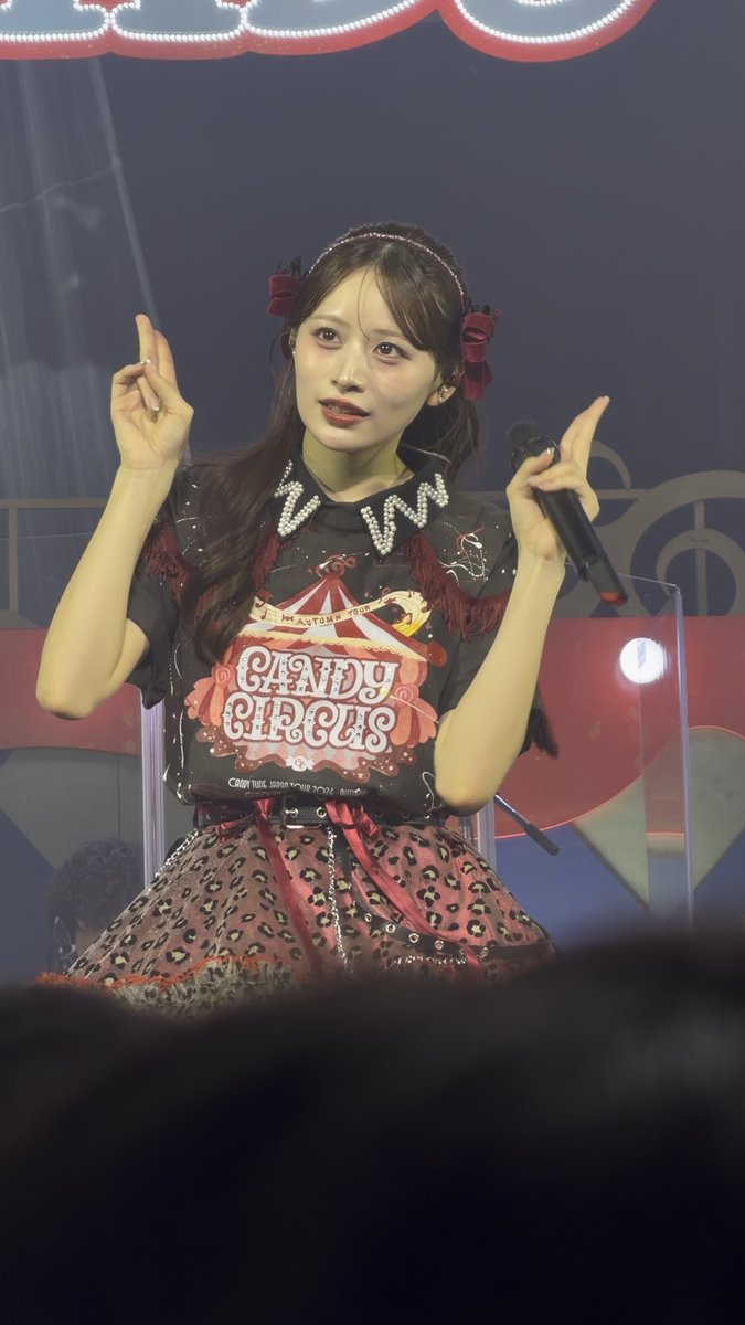 A person on stage wearing a black t-shirt with 'CANDY CIRCUS' printed on it, a black and pink leopard print skirt, and a black headband with roses. They are holding a microphone and have their finger pointed up.