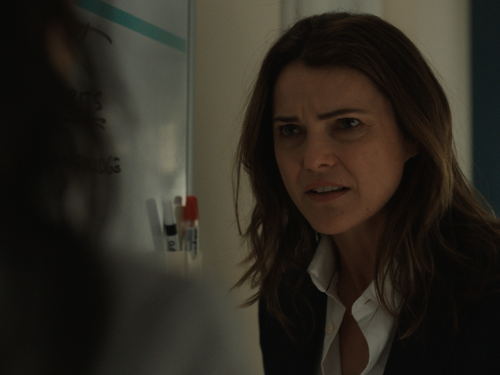 A scene from The Diplomat featuring Keri Russell as Kate Wyler, her face furrowed with concern as she appears to be in an intense conversation, with marker pens and writing visible on a whiteboard behind her.