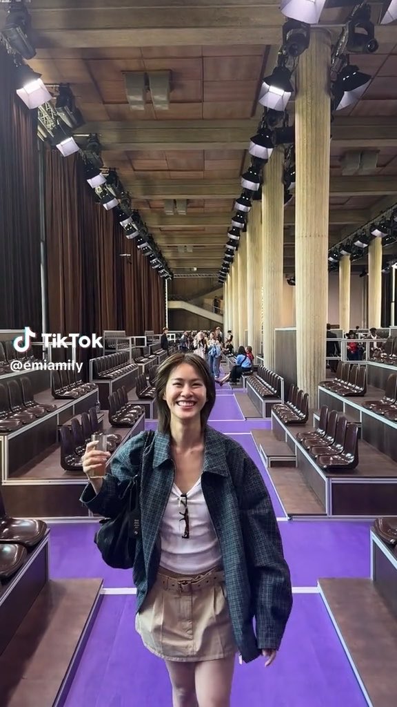 A TikTok screenshot by @emiamily showing a smiling young woman in a green plaid jacket and shorts posing in what appears to be a large venue with rows of seats and extensive lighting equipment overhead.