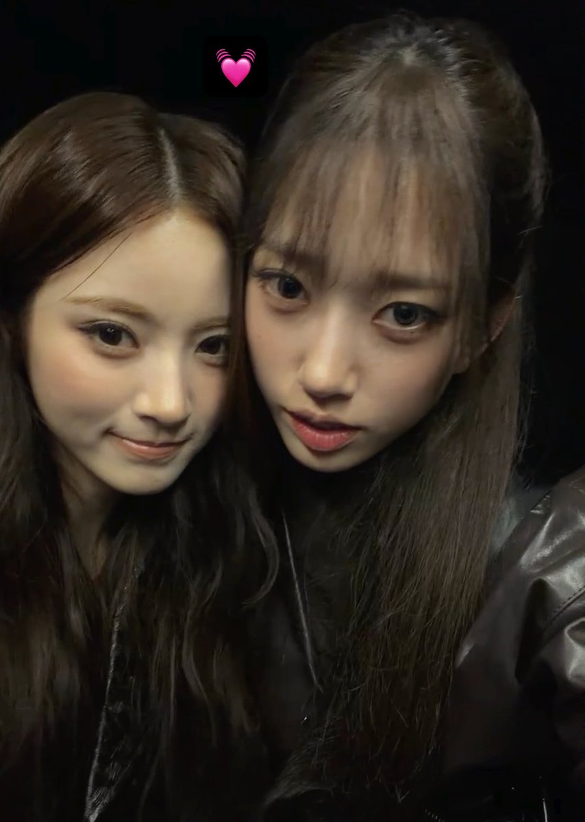 Two young women with long dark hair pose closely together for a selfie in low lighting, with a pink heart-with-soundwaves emoji emoji placed above them.