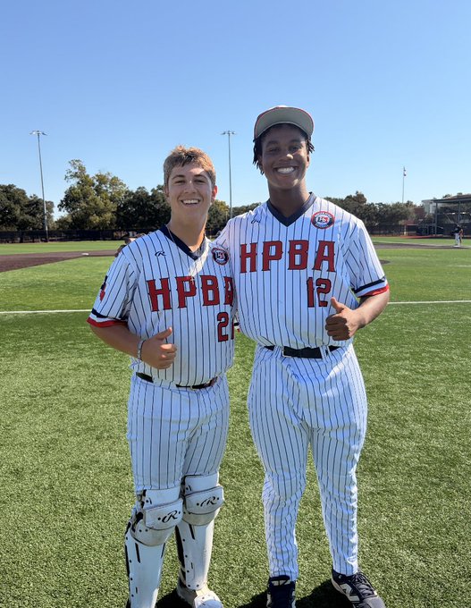 Two young baseball players in white pinstriped HPBA uniforms standing on a baseball field, both smiling and giving thumbs up.