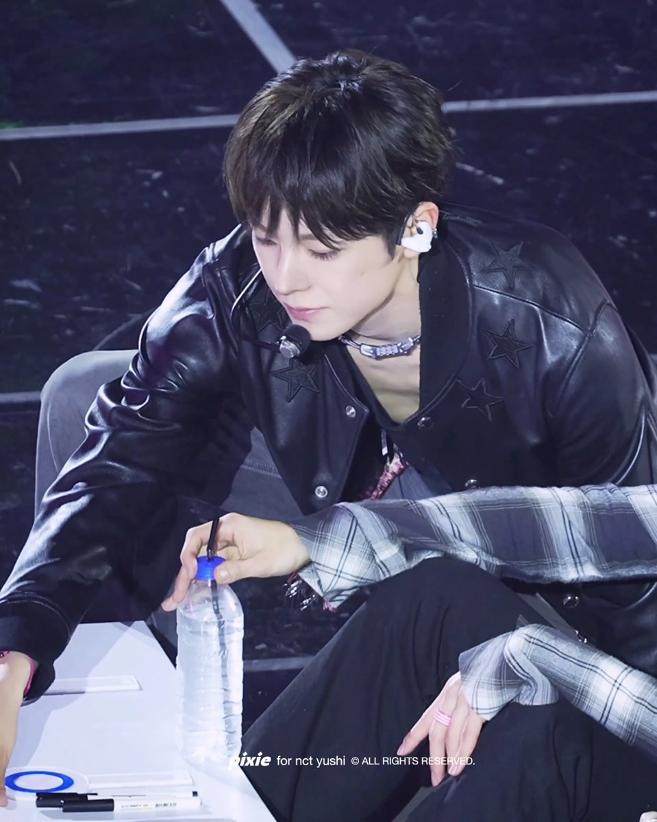 A young man with dark hair in a black star-patterned leather jacket looks down while holding a water bottle, with a watermark reading 'pixie for nct yushi © ALL RIGHTS RESERVED.'
