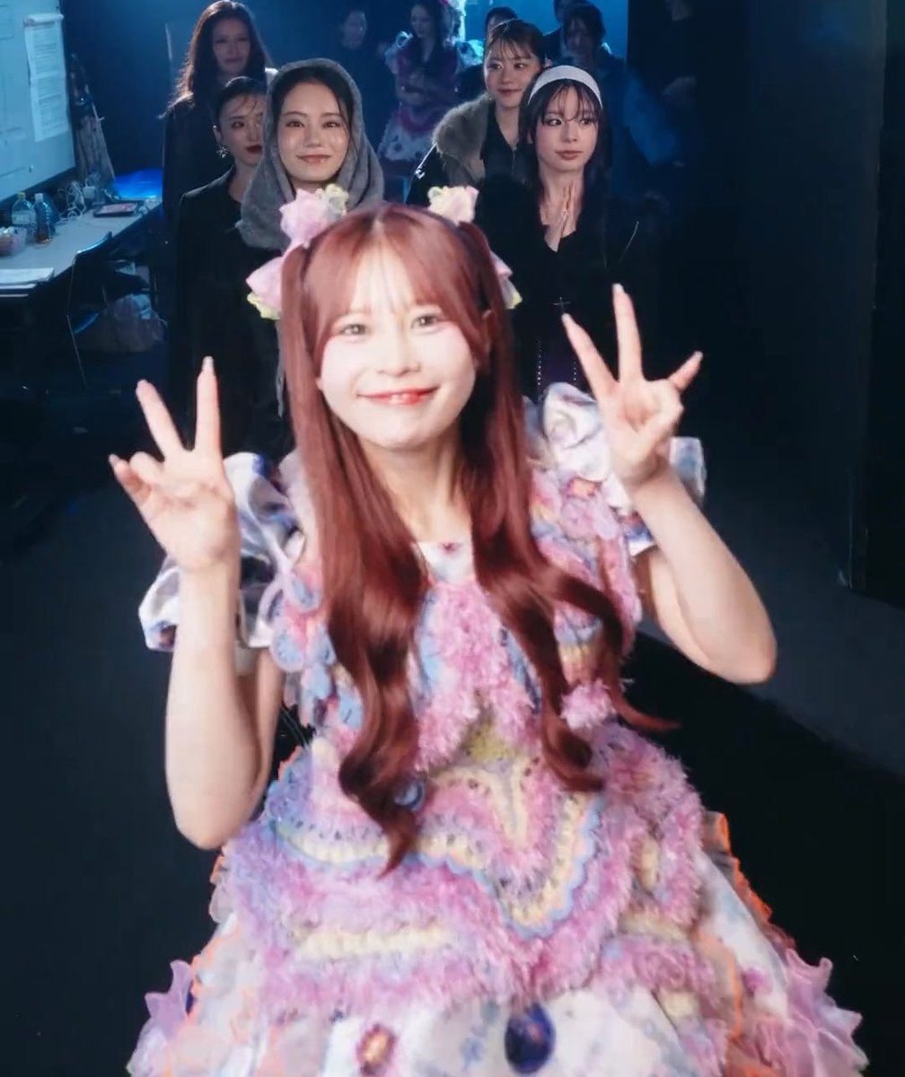 A photo of a smiling woman with long reddish-brown hair in a frilly pastel costume making peace signs with both hands, surrounded by other women backstage.