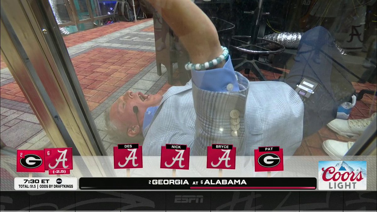 A man in a checkered blazer and blue shirt is shown lying on his back on a "College Gameday" set, appearing to laugh as he looks up at someone standing over him. The ESPN ticker displays "Georgia at Alabama" with team members Des, Nick, Bryce, and Pat pictured, alongside a Coors Light logo, capturing the moment referenced in the caption.