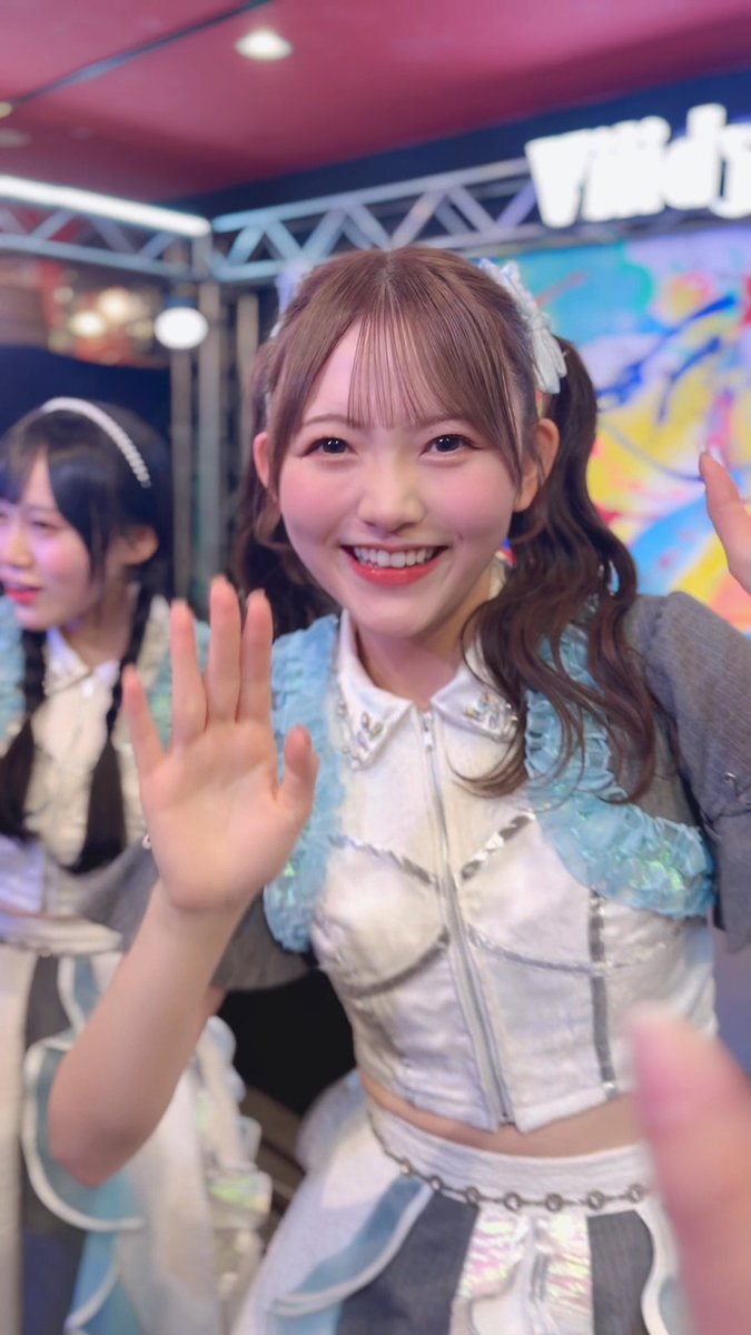 An idol performer in a blue and white outfit waves enthusiastically at the camera.