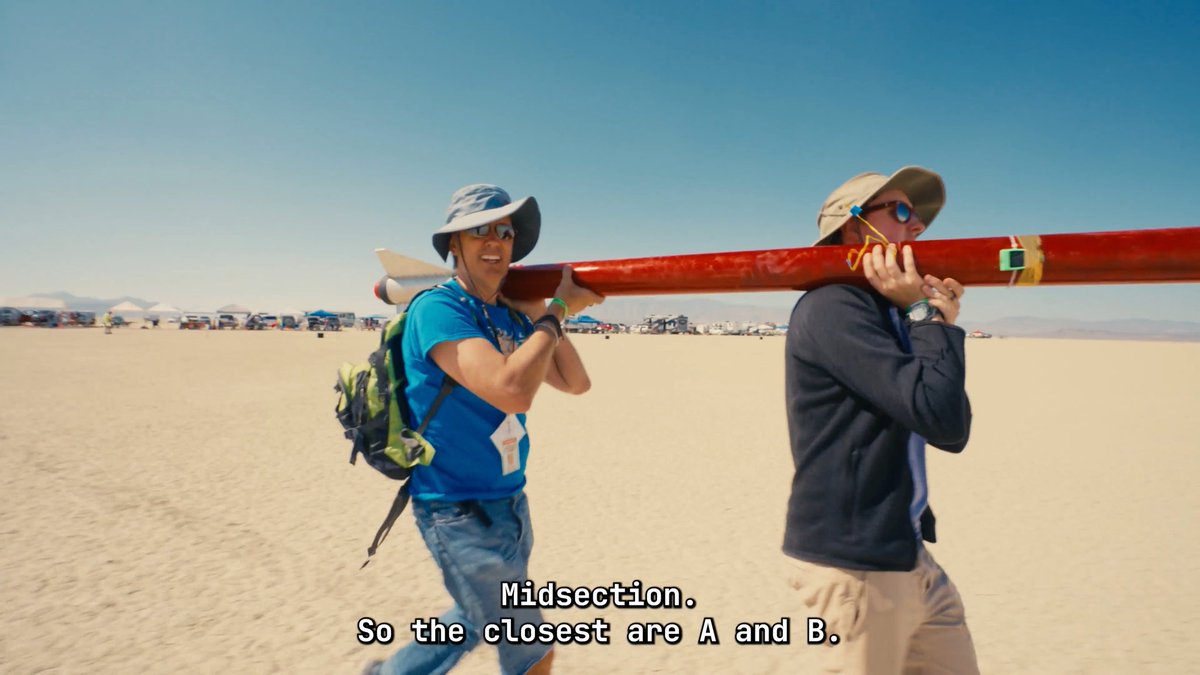 Two people in a desert landscape, one wearing a blue shirt and bucket hat, the other in a dark hoodie, carrying a large red model rocket on their shoulders, with subtitles reading 'Midsection. So the closest are A and B.'
