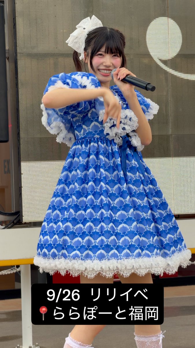 A smiling young woman in a blue and white scalloped-pattern stage dress performs on stage holding a microphone, with Japanese text reading "9/26 リリイベ 📍ららぽーと福岡" (Release event at LaLaport Fukuoka on 9/26).