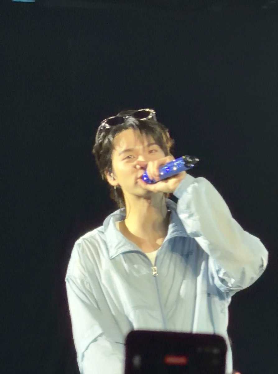 A male performer wearing a light blue jacket and sunglasses on his head sings into a blue microphone on stage.