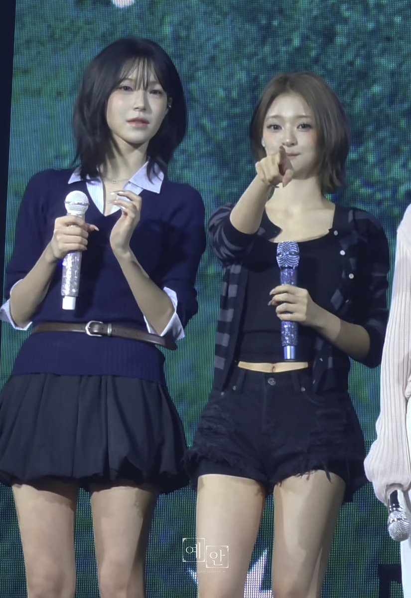 Two young women, likely K-pop performers, on stage holding sparkling microphones; one wears a navy sweater with a white collar and dark skirt, while the other wears a black top and frayed black shorts.