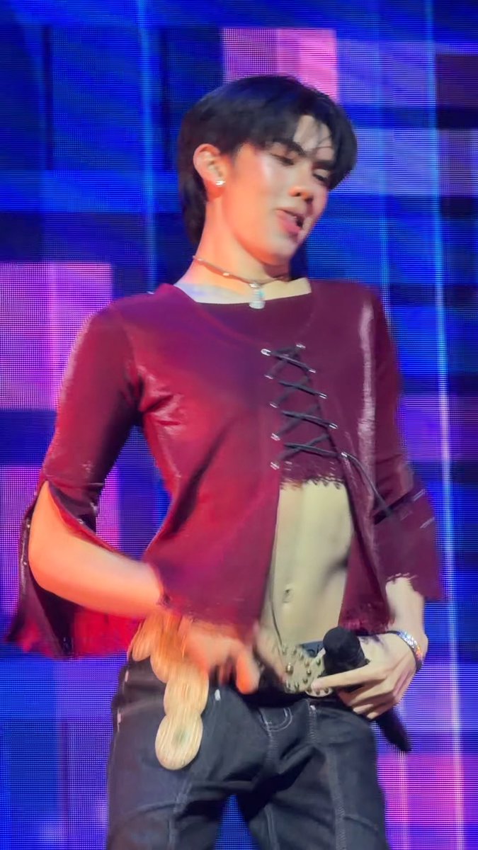 A performer on stage wears a burgundy lace-up top and dark jeans, holding a microphone with a decorative gold piece, set against a purple-lit background.