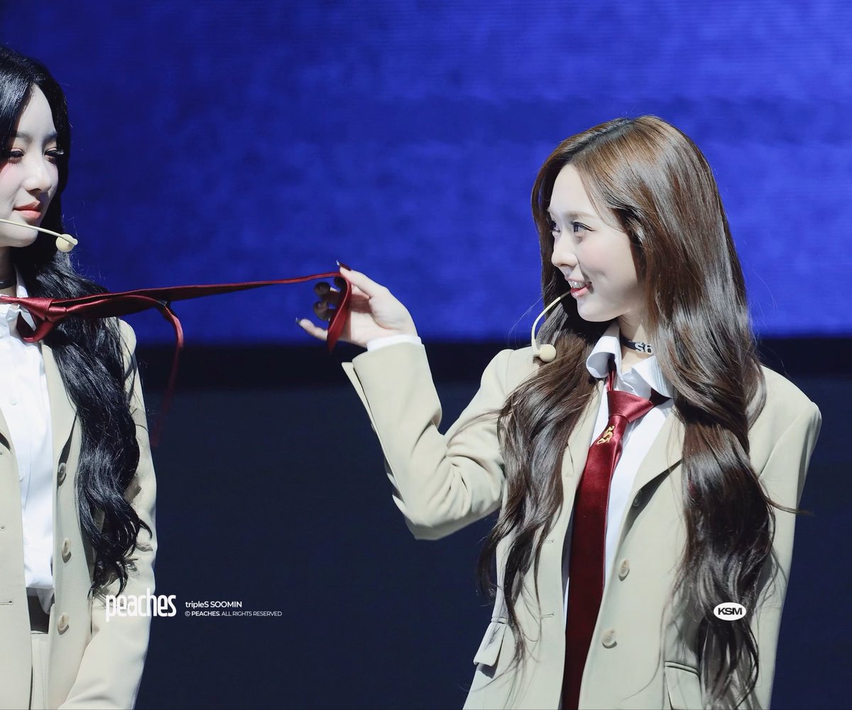 Two members of tripleS, identified by the watermark as Soomin, wearing matching beige blazers and white shirts, playfully holding a red tie; the image is watermarked "peaches tripleS SOOMIN" and "KSM."