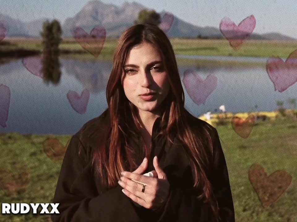 A young woman with long reddish-brown hair in a black shirt stands outdoors with hands clasped, with semi-transparent heart overlays floating across the image; the watermark 'RUDYXX' appears in the bottom left.