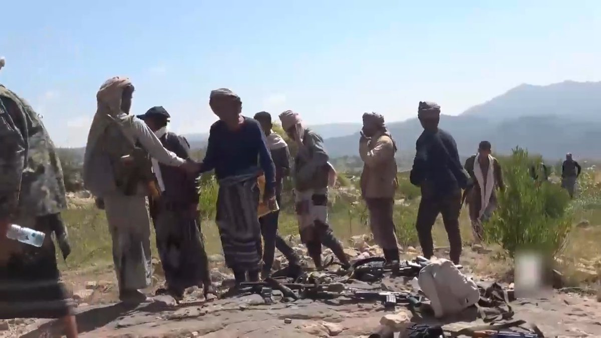 A group of men in traditional Middle Eastern attire stand outdoors in a rocky, mountainous landscape with weapons and equipment scattered on the ground in front of them.