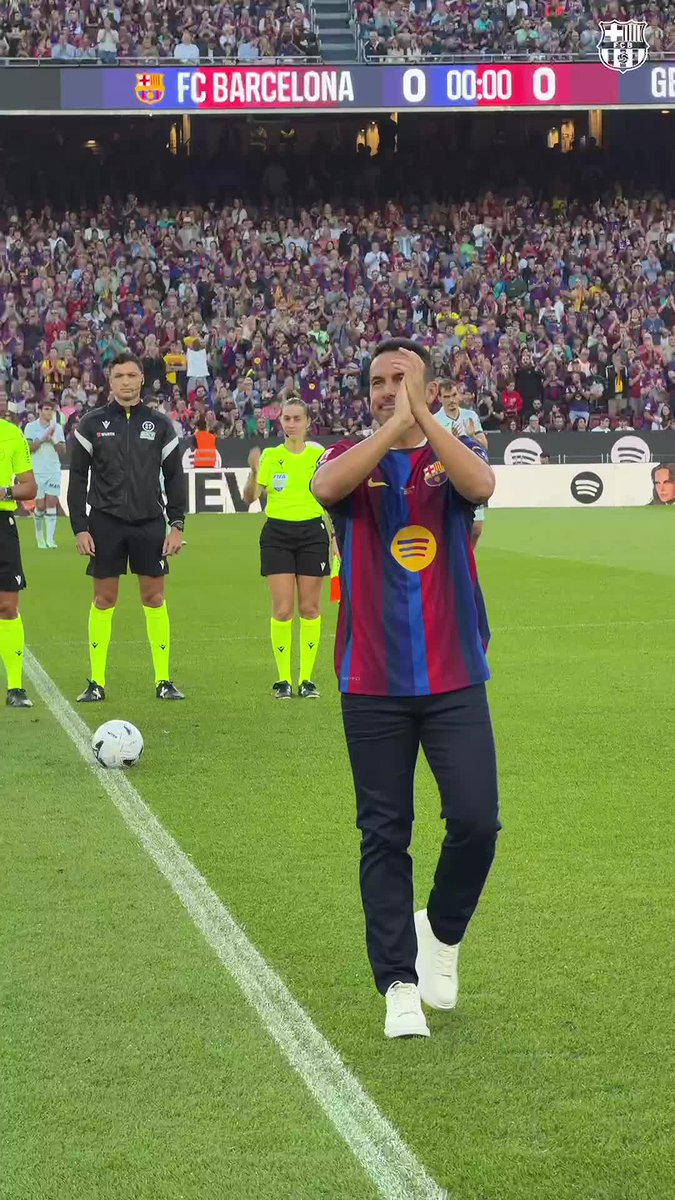 Barcelona legend Pedro Rodríguez, wearing the iconic blue and red Barça jersey, applauds the crowd during an honorary kick-off ceremony at a packed Camp Nou, with the scoreboard showing "FC BARCELONA 0 00:00 0" before the match begins.