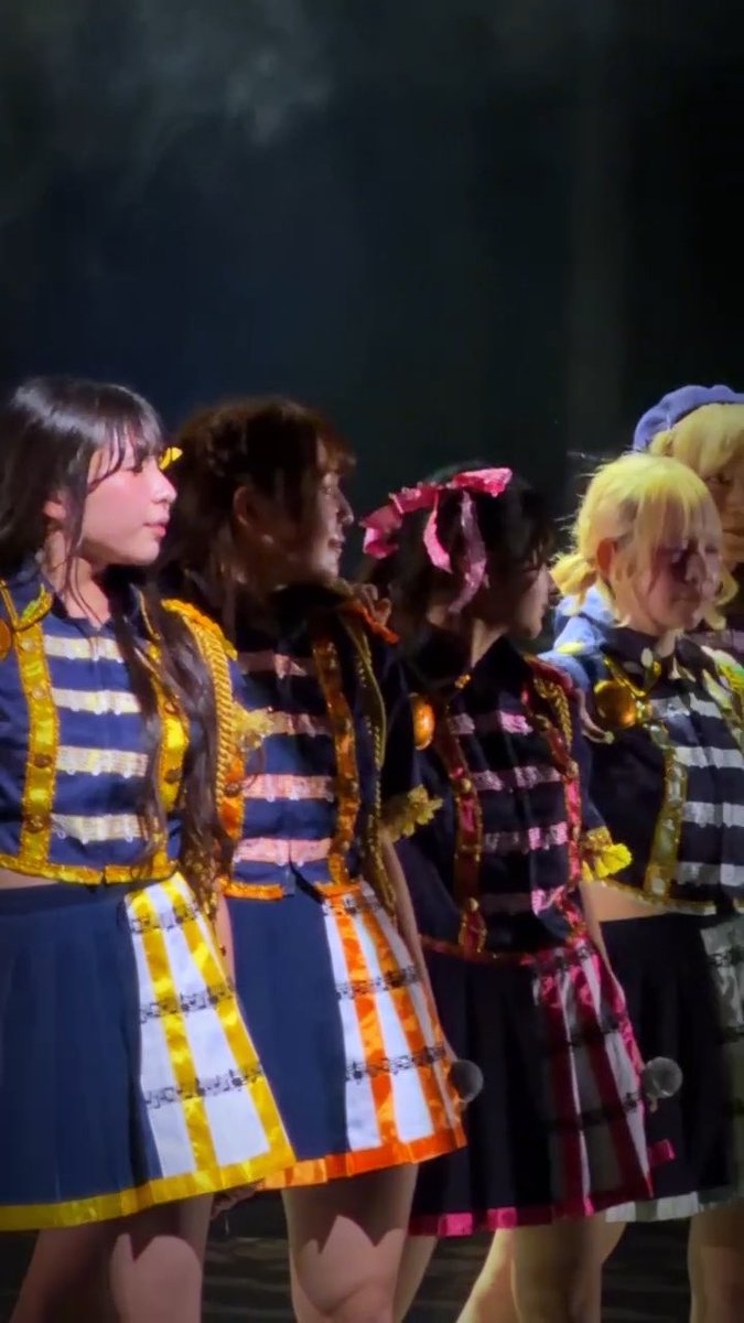 A group of female performers in matching dark blue, gold, and pink military-style costumes stand in a line on a dimly lit stage.