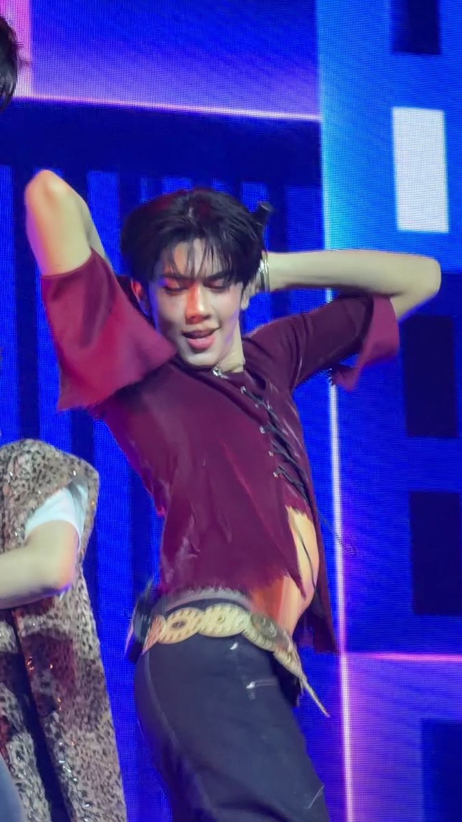 A young male performer in a distressed maroon shirt and dark pants dances on stage under blue lighting, with his hands behind his head.