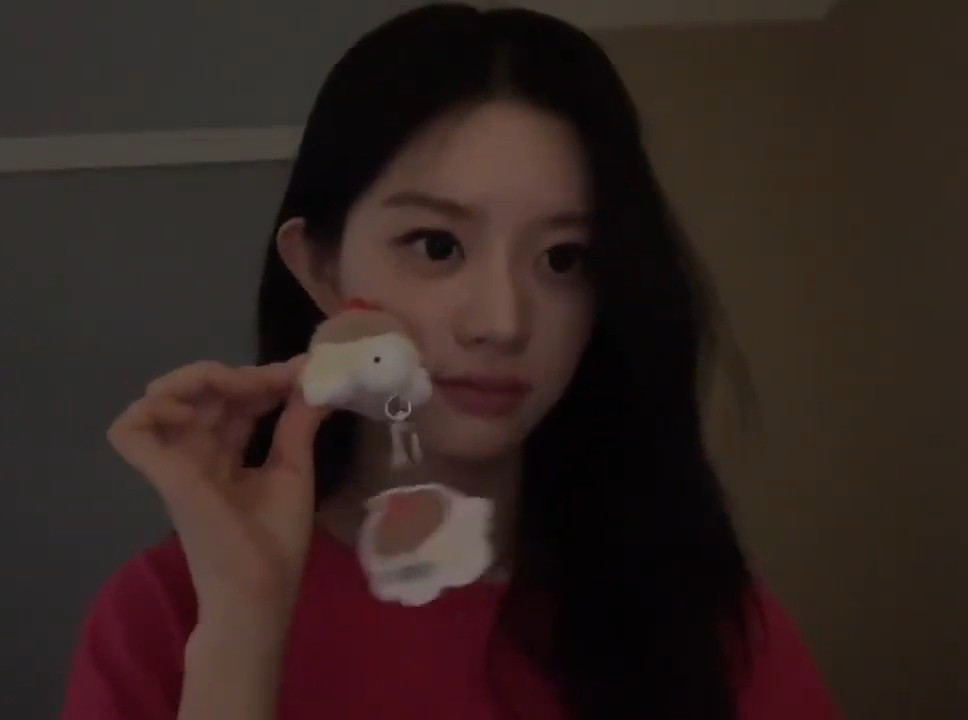 A young woman with long dark hair, wearing a pink shirt, holds up a small, fluffy white bunny-shaped keychain charm.