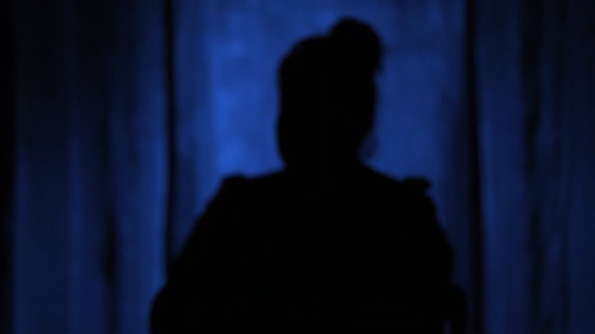 A dark silhouette of a person standing against a deep blue illuminated background, possibly in a forest setting.